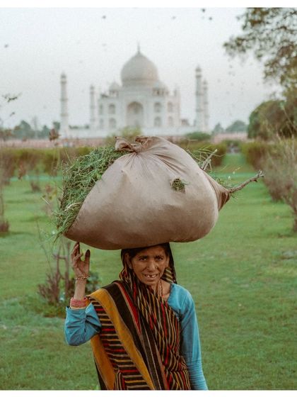A woman carrying a large bundle of grass on her head pauses for a portrait in front of the Taj Mahal. Her strength and resilience are as monumental as the building behind her.