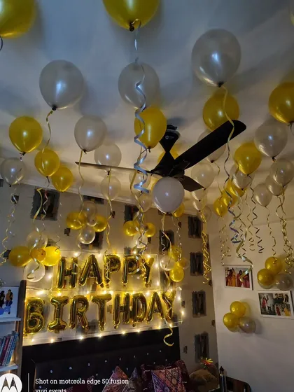 A cheerful birthday room decoration using yellow and white balloons floating on the ceiling. The 'Happy Birthday' foil letters are highlighted with warm fairy lights, creating a bright and happy vibe.