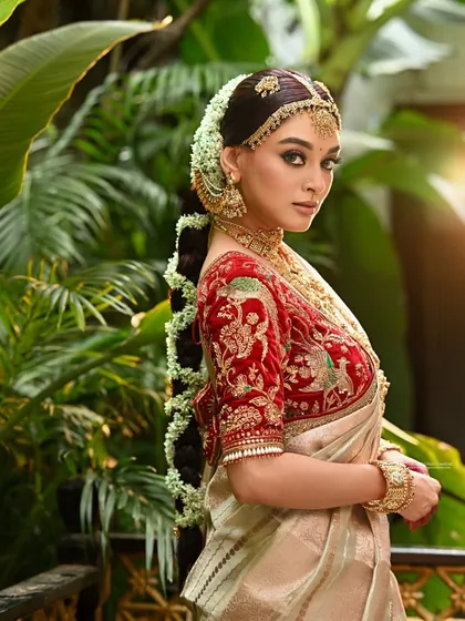 A side profile of a model in a South Indian bridal look. This angle beautifully showcases the elaborate hairstyle with fresh flowers, the detailed embroidery on the blouse, and the layered gold jewellery.