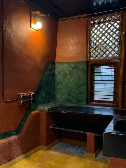 A kitchen in a mud cottage, with a green oxide backsplash and a layered window design featuring bamboo lattice for ventilation.