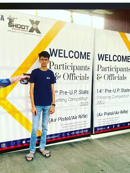 A participant stands before the official banner for the 14th Pre-U.P. State Shooting Competition, an event we were proud to organize at Ramagya School. We are dedicated to promoting the sport of shooting at all levels.