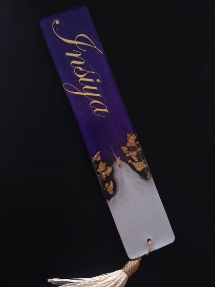A deep purple and white resin bookmark made for 'Insiya', with the name in gold script and gold foil accents. The tassel is a complementary cream color.
