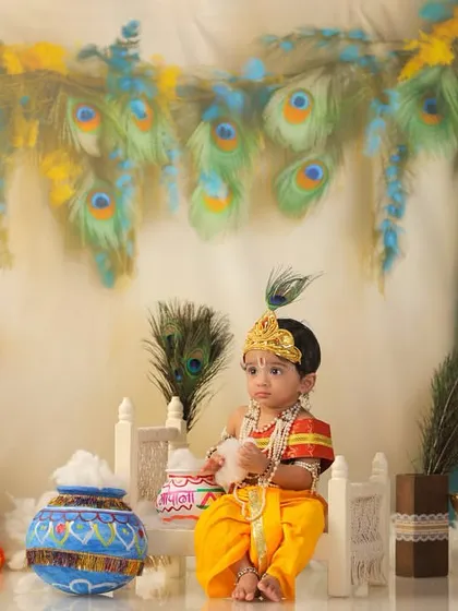 A full portrait of a baby boy dressed as Lord Krishna, seated amidst a divine setup with peacock feathers, a flute, and traditional matkis (pots).