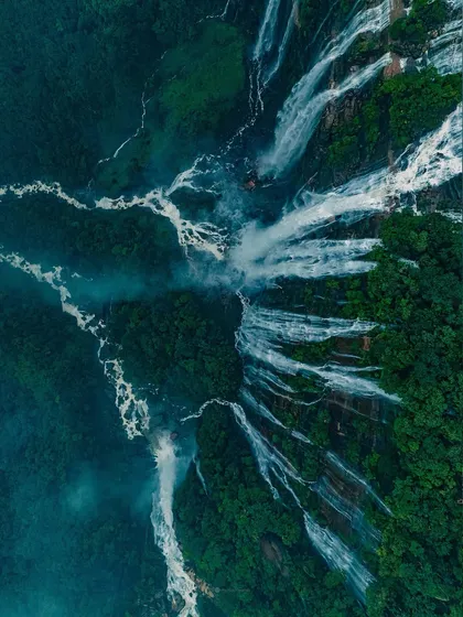 A dramatic top-down drone shot of waterfalls in Meghalaya, showing the raw power of water as it carves through the dense green forest.