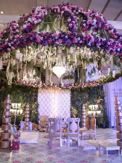 A breathtaking floral mandap, with a dense canopy of purple and pink flowers and hanging chandeliers, creating an enchanted garden atmosphere for the wedding ceremony.