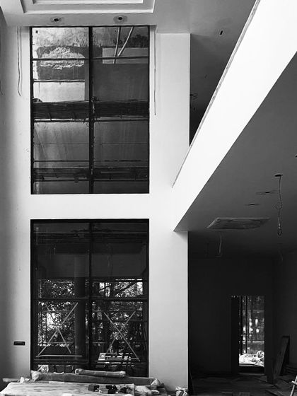 An interior view of a double-height room under construction, focusing on the dramatic play of light and shadow created by the large, unfinished window frames.