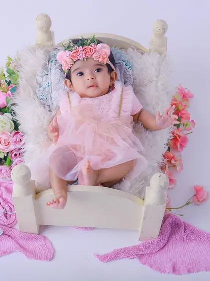A sweet baby girl in a pink dress and floral headband, resting on a miniature bed. The soft textures and pastel colors create a delicate and feminine portrait.