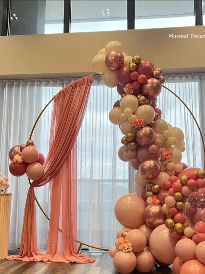 This elegant balloon arrangement features a large golden ring draped with pink fabric. The organic garland of pastel pink, cream, and rose gold balloons creates a soft and romantic backdrop.