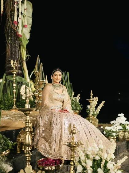 A stunning portrait of the bride seated at the traditionally decorated mandap. The combination of brass elements, white florals, and her golden lehenga creates a timeless and regal image.