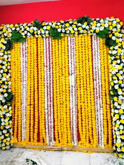 This vibrant floral wall was designed for a Mehndi ceremony at the bride's home. It features a dense curtain of yellow and white marigold strings, framed by a border of white and yellow chrysanthemums and fresh green leaves.