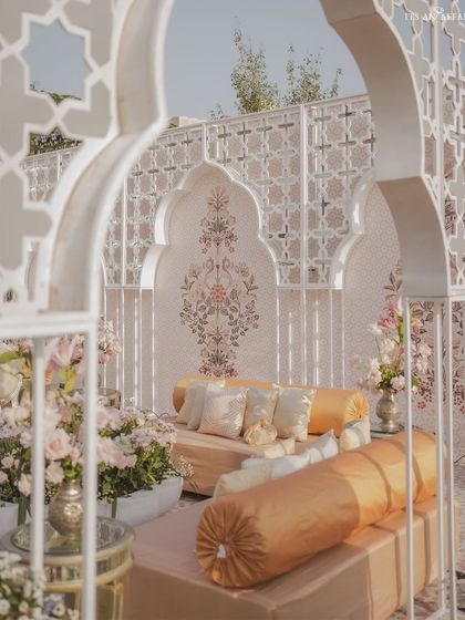 A cozy seating nook with traditional gaddas and bolsters in shades of cream and gold. The backdrop features a hand-painted motif, viewed through an elegant jali archway.