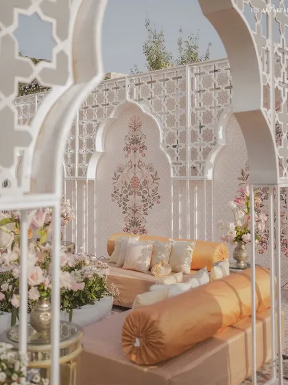 A cozy seating nook with traditional gaddas and bolsters in shades of cream and gold. The backdrop features a hand-painted motif, viewed through an elegant jali archway.