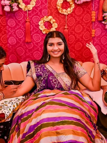 The bride getting her henna done, seated against a backdrop of bright pink fabric and traditional 'gota patti' floral wreaths.