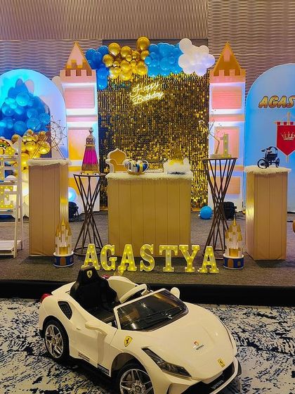 We can create a royal celebration with a castle theme. This setup for Agastya features castle turrets, a golden sequin backdrop, and props like a toy horse and a miniature Ferrari for the little prince.