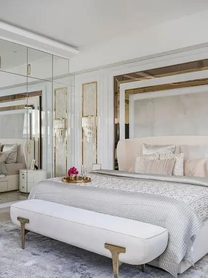 In this master bedroom, we used mirrored wardrobe doors to create a sense of spaciousness. The bed is framed by layered paneling with gold accents, and the all-white furniture and soft furnishings create a serene and luminous feel.