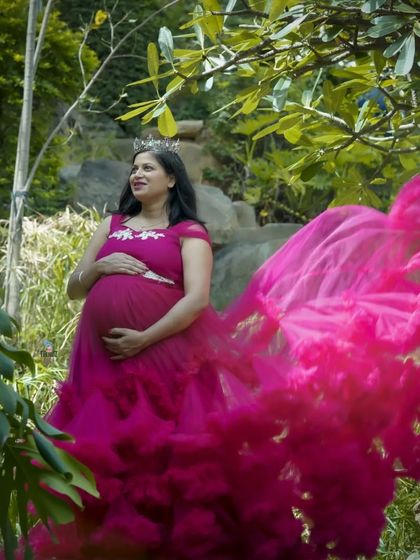 A regal solo portrait in a lush garden setting. The mom-to-be wears a small crown, adding a touch of royalty to this beautiful shot in a flowing magenta gown.