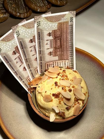 A playful and creative dessert presentation. This rich, nutty kulfi is served with edible currency notes, adding a fun and memorable element to the dining experience.