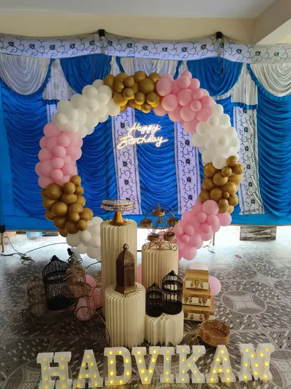 This circular balloon arch in pink, white, and gold is perfect for outdoor or tented events. The design includes a 'Happy Birthday' neon sign and the name in light-up letters, ensuring the decor stands out.