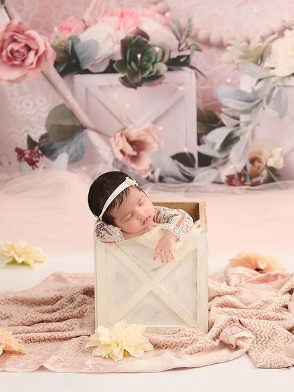 A baby girl peeks out of a white wooden box in a beautifully styled pink and white set with floral crates and soft fabrics.