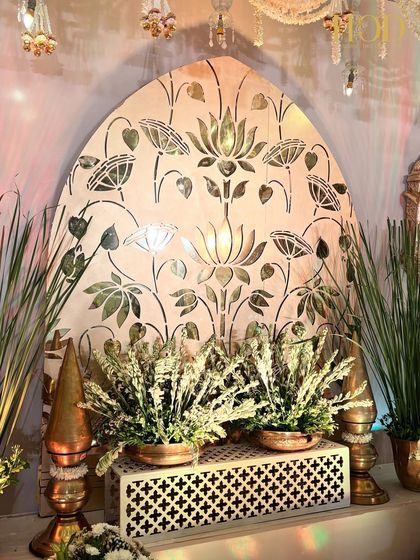 A beautiful setup featuring a white jali panel with a lotus cutout, in front of which are placed traditional brass urlis and floral arrangements.