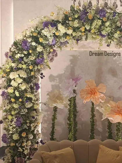 A close-up of a beautiful floral arch designed for a baby shower. The mix of white, yellow, and purple flowers is complemented by whimsical, oversized paper flowers in the background.