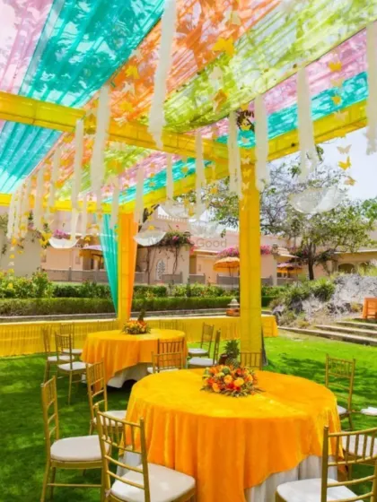 An outdoor daytime Mehendi or Haldi lunch setup. The colorful canopy provides shade while adding to the festive look, with bright yellow tablecloths completing the theme.