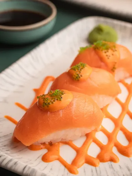 A close-up of salmon nigiri, beautifully plated with a spicy mayo drizzle. The tight crop focuses on the freshness of the fish and the vibrant colors.