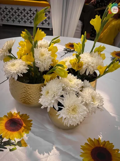 A bright and sunny table centerpiece for a Haldi, with yellow and white flowers in textured golden pots on a sunflower-print tablecloth.