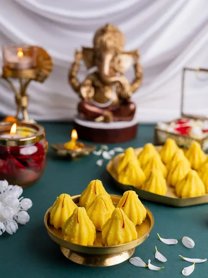 A beautiful arrangement of Kesar Modaks for a festive offering. The soft lighting and scattered petals create a serene and divine mood, with the golden Ganesha idol adding a spiritual touch.