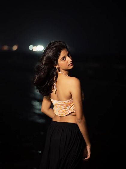 A night portrait taken at the beach, with the distant city lights creating a soft bokeh. The wind in her hair adds a natural, dynamic element to the shot.