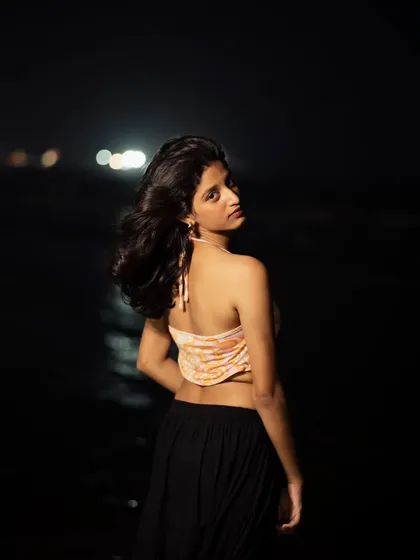 A night portrait taken at the beach, with the distant city lights creating a soft bokeh. The wind in her hair adds a natural, dynamic element to the shot.