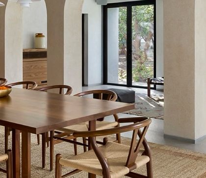 The dining area in the Mango Orchard House is defined by elegant archways and a long wooden table. The wishbone-style chairs add a touch of classic mid-century design.