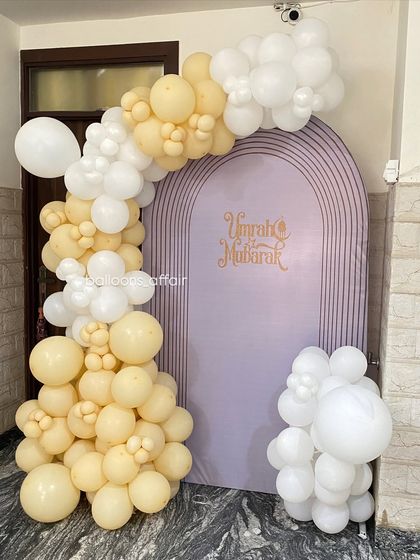 An elegant 'Umrah Mubarak' decoration to welcome loved ones. The setup features a lavender arched backdrop with a beautiful balloon garland in pastel yellow and white.
