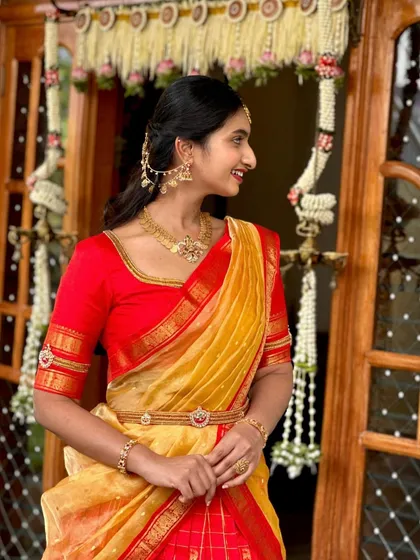 A side profile of the client, highlighting the elegant drape of the yellow organza dupatta and the intricate kasu work on the red silk blouse sleeves.