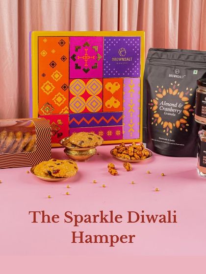 The Sparkle Diwali Hamper is all about festive joy. This beautifully styled shot shows the vibrant packaging alongside the contents, including our Almond & Cranberry Granola, assorted cookies, and jars of spiced nuts and seeds.