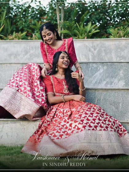 Two friends celebrating together in our bespoke red and pink Banarasi lehengas, showcasing different color variations of the same design.
