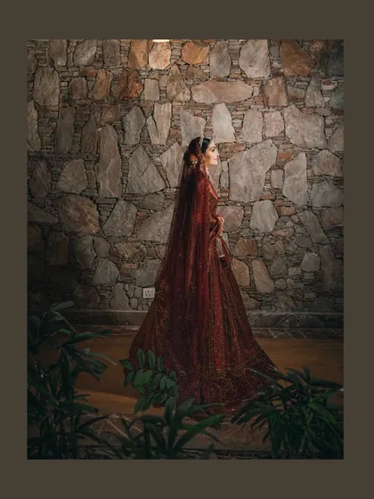 A stunning portrait of the bride in her deep maroon lehenga, her silhouette beautifully captured against a stone wall.