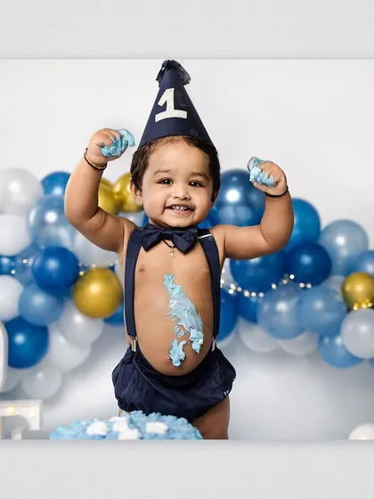 A triumphant birthday boy raises his frosting-covered hands in the air, celebrating his first birthday with a cake smash. The blue and gold balloon arch adds a festive touch to the studio session.