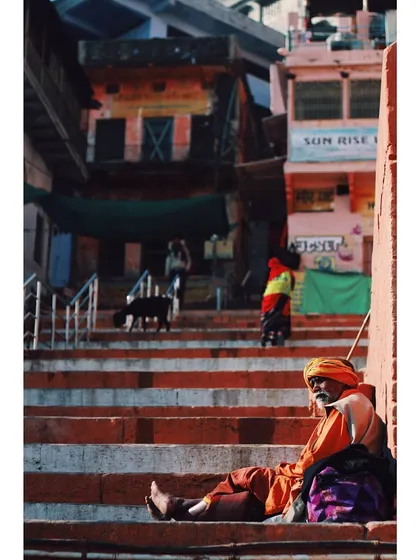 A sadhu rests on the steps of a ghat, bathed in the warm morning light. The vertical composition emphasizes the layers of life happening on the ghats, from the resting man to the people in the background.