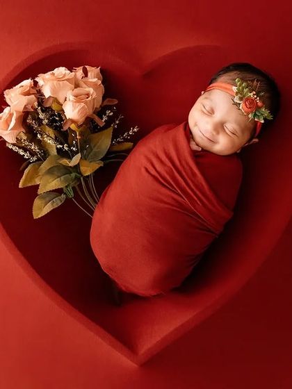 A smiling newborn swaddled in red rests in a heart bowl with a bouquet of roses, a joyful and vibrant portrait.