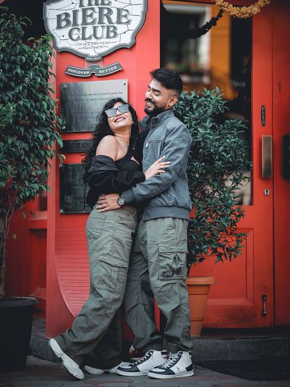 A fun and stylish shot of a couple in matching cargo pants, embracing in front of the iconic red entrance of 'The Biere Club' in Bangalore.