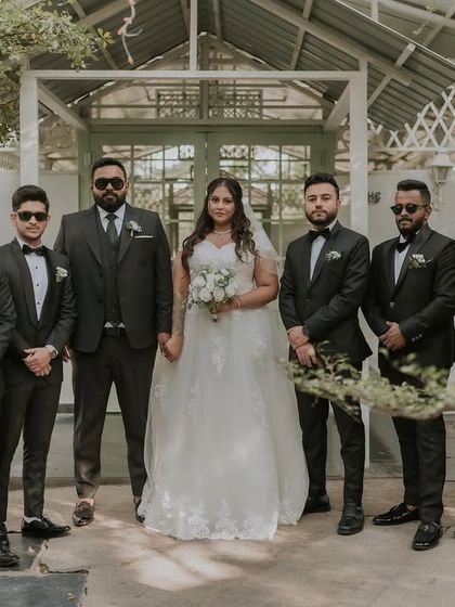 A bride and her groomsmen pose for a classic group photo. The clean, white architecture of the cottage provides a great contrasting background for their dark suits.