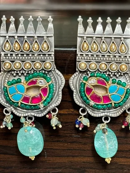 A vibrant pair of fusion earrings. These feature a dual-tone design with colourful enamel work in a peacock motif, finished with dangling light blue gemstone drops.