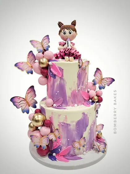 A two-tier butterfly-themed cake with a beautiful purple and pink watercolor effect, adorned with a cute girl figurine.