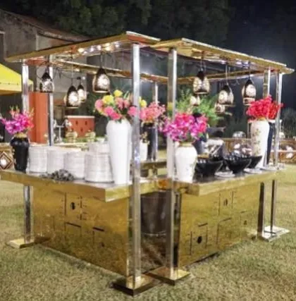 A unique, golden, four-sided catering stall designed for outdoor events. This island setup is decorated with hanging lamps and colorful flowers, creating a beautiful and accessible food station from all sides.