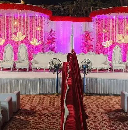 A grand wedding stage with a traditional feel. The setup features ornate seating, hanging floral decorations, and dramatic pink and red lighting.