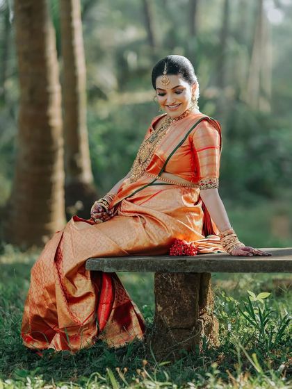 A serene moment captured outdoors. The rich red of the Kanchipuram silk saree contrasts beautifully with the tranquil nature setting.
