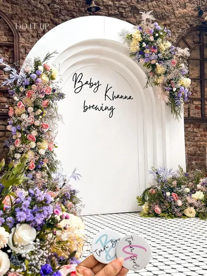 A stunning floral archway for a baby shower. The white arched backdrop, which reads 'Baby Khanna brewing', is adorned with an abundance of pastel flowers, creating a perfect photo opportunity and entrance piece.