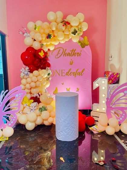 A "one-derful" butterfly-themed first birthday decoration. The setup uses a pink arch backdrop, a pastel yellow and red balloon garland, and large butterfly wing cutouts next to a light-up number '1'.