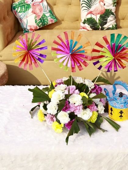 Even the seating areas get a touch of personality. We created this cozy corner with fun, tropical-print cushions and a centerpiece featuring colorful pinwheels and a quirky painted kettle, perfect for a relaxed Mehendi vibe.